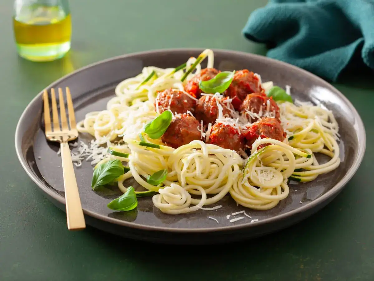 Pyszna keto kolacja: klopsiki w sosie pomidorowym z makaronem z cukinii, posypane parmezanem i listkami bazylii. Złoty widelec czeka.
