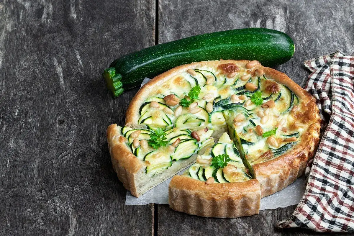 Torta salata senza uova con zucchine, pinoli e prezzemolo su un tavolo di legno scuro.