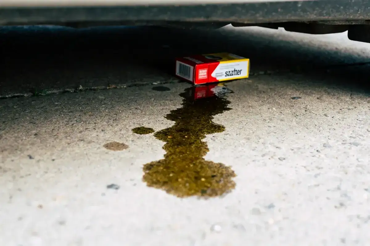 Une flaque d'huile sous une voiture, signe d'une fuite joint spi bo&icirc;te de vitesse. Une petite bo&icirc;te rouge et jaune est pos&eacute;e &agrave; proximit&eacute;.