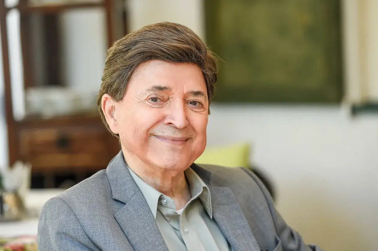 Uśmiechnięty Jerzy Połomski w szarym garniturze. W tle widać meble i zieloną ścianę.
