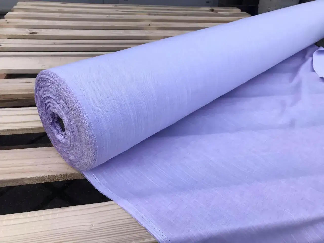Jasnofioletowy materiał o splocie **twill** nawinięty na rolkę, leżący na drewnianych paletach.