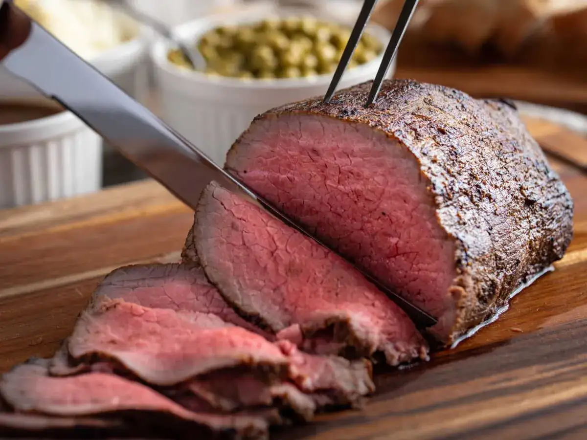Un rosbeef juteux est tranch&eacute; sur une planche en bois. Le temps de cuisson rosbeef au four est parfait pour cette viande ros&eacute;e.