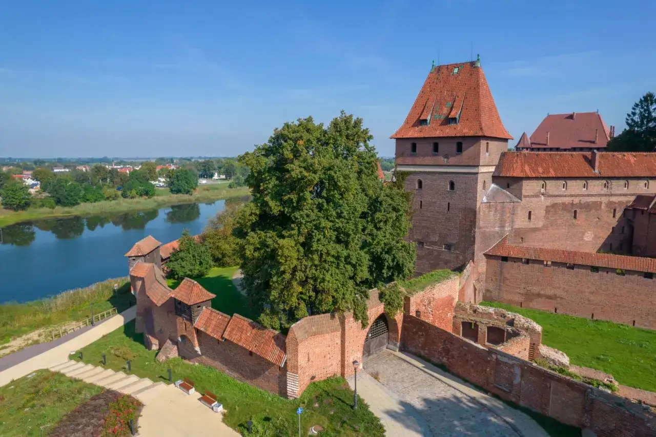 Potężna ceglana twierdza, z charakterystyczną czerwoną jesionką na dachu, góruje nad spokojną rzeką i zielonymi terenami.