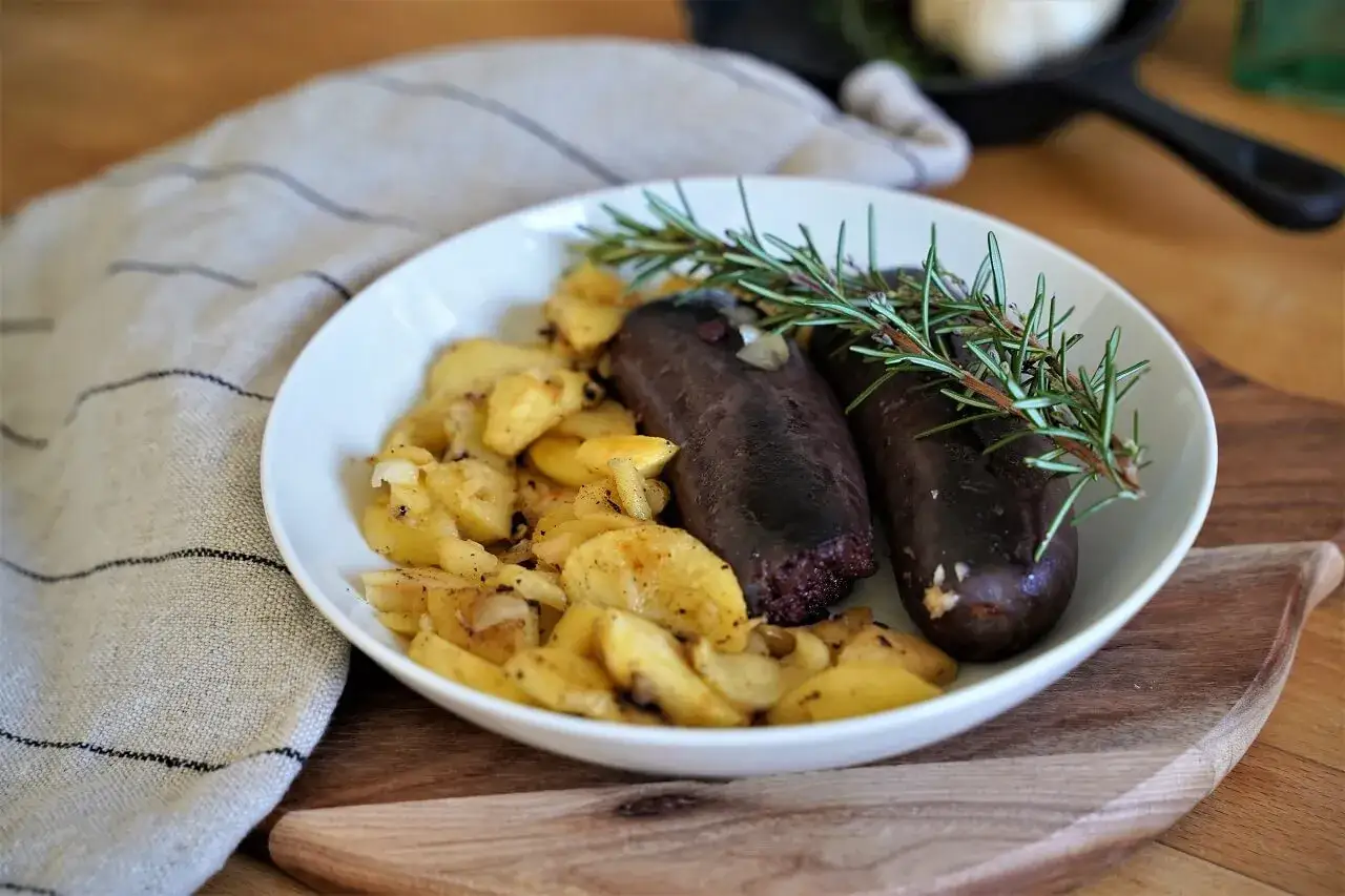 Deux boudins noirs savoureux, accompagnés de pommes de terre sautées et d'une branche de romarin. La cuisson boudin noir est mise en valeur dans cette assiette appétissante.