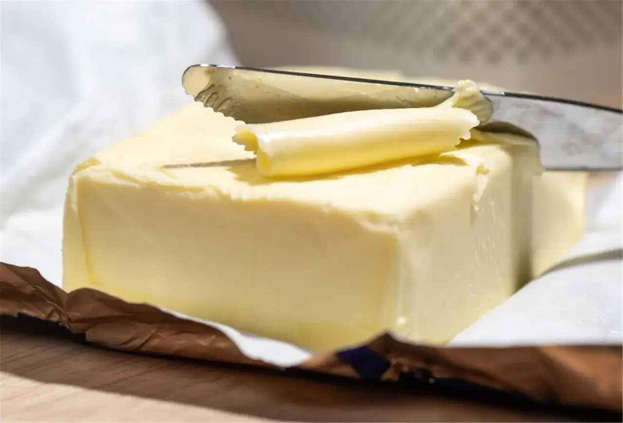 Ein Messer schneidet eine Scheibe von einem Block Butter ab. Die goldgelbe Butter liegt auf ihrer Verpackung.