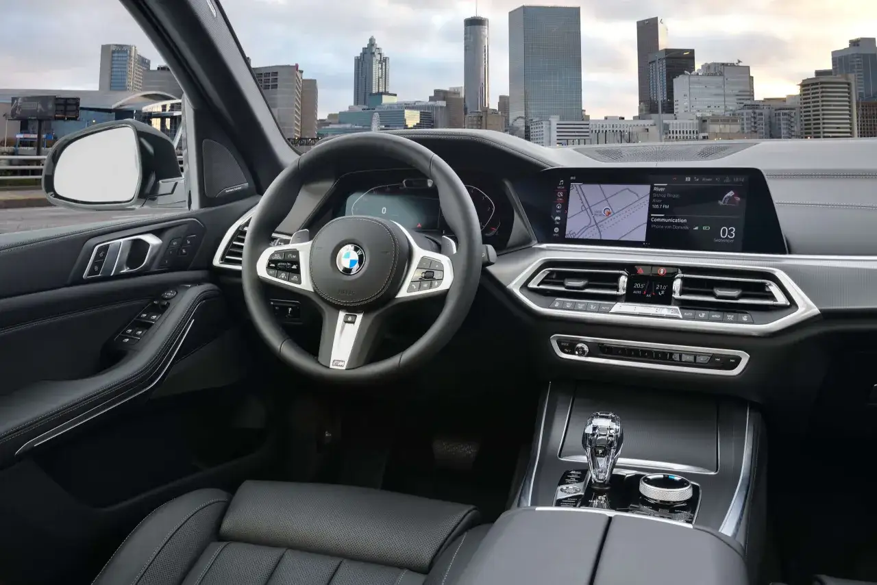 Wnętrze BMW X5, widok na kierownicę, ekran multimediów z nawigacją i miasto w tle. Informacje z BMW X5 wiki.