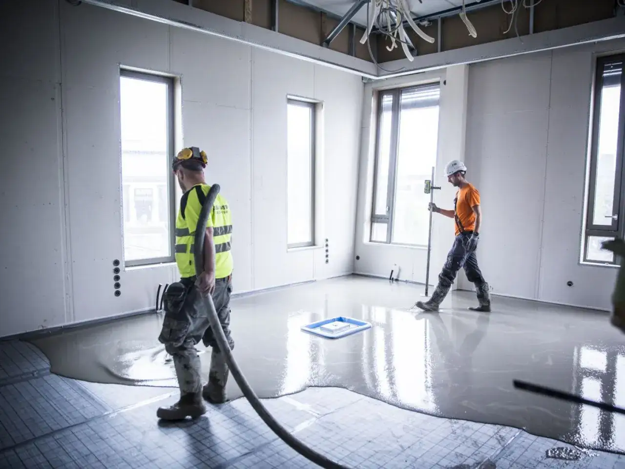 Dwóch budowlańców pracuje nad wylewką samopoziomującą. Jeden z nich kieruje wylewkę wężem, drugi sprawdza poziom.