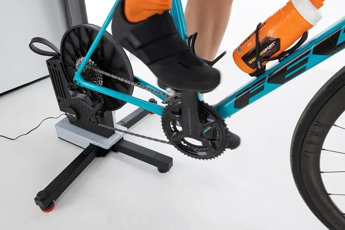 Rowerzysta trenuje na trenażerze z napędem **direct drive**. Widoczna jest jego stopa w czarnym bucie kolarskim, korba i łańcuch.