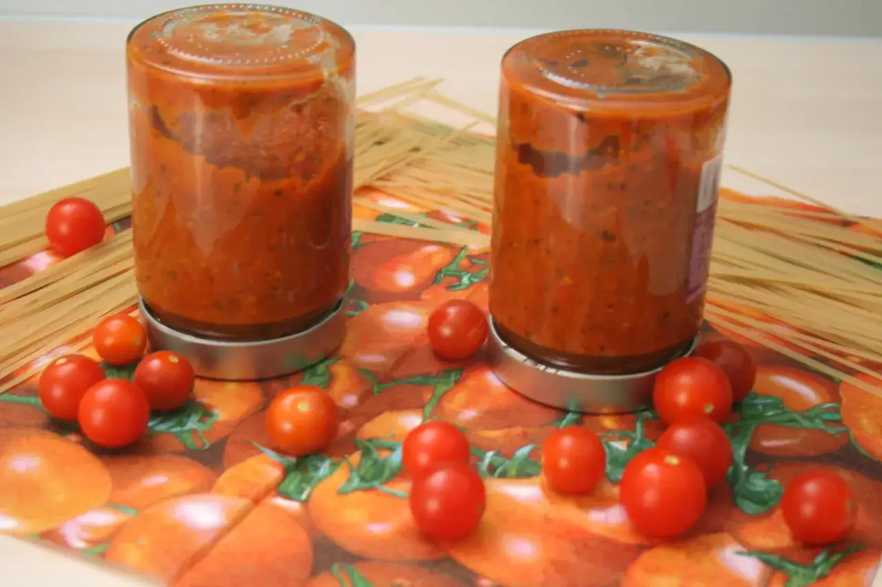 Zwei Gl&auml;ser hausgemachtes tomaten sugo stehen neben Spaghetti und frischen Kirschtomaten auf einer Serviette mit Tomatenmotiv.