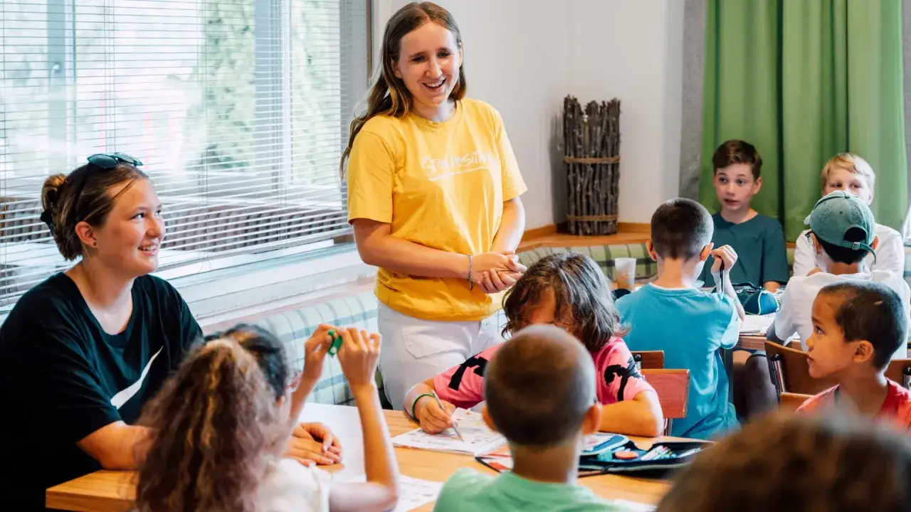 Lachende Betreuerin leitet ein englisch camp. Kinder lernen und malen an Tischen.