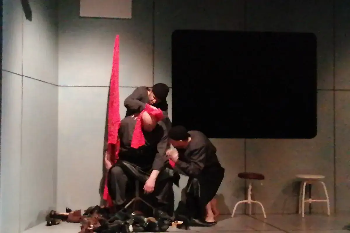Na scenie mężczyźni w czerni, z czerwonymi chustami, tworzą intymną scenę. Ich gesty przypominają fragment erotycznego opowiadania o stopach.