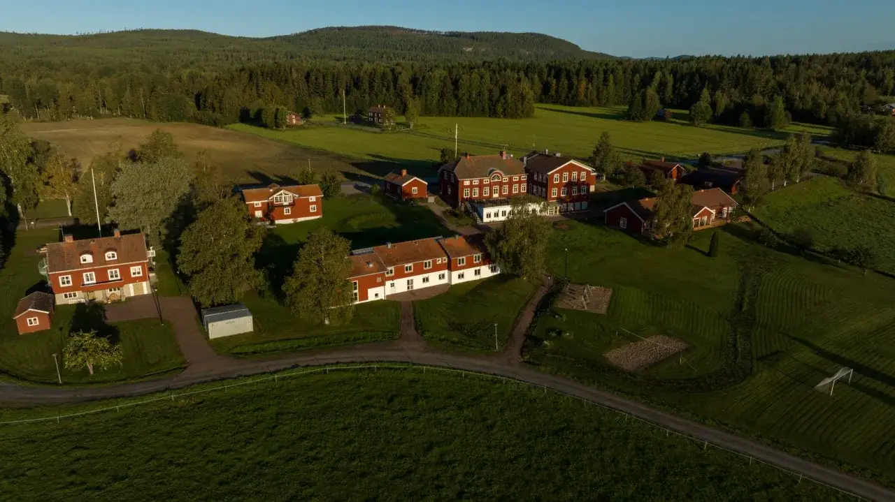STF Undersvik, en idyllisk anl&auml;ggning med r&ouml;da tr&auml;hus och gr&ouml;nskande f&auml;lt, omgiven av skog och en klarbl&aring; himmel.
