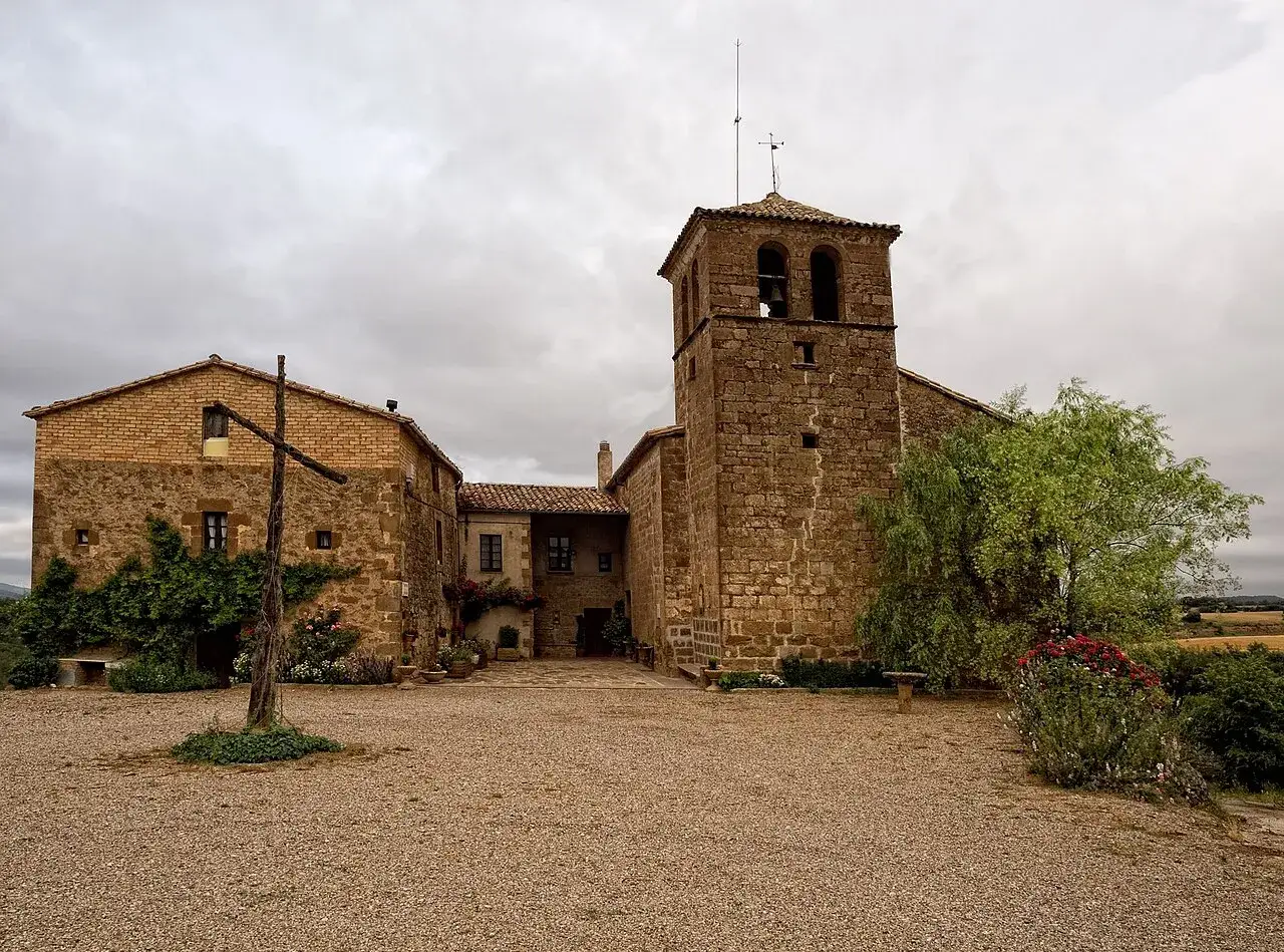 Antigua ermita de piedra en Pinell de Solson&egrave;s, con un pozo de madera y vegetaci&oacute;n exuberante.
