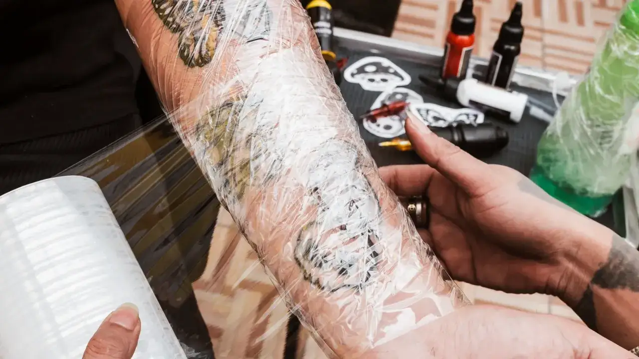 Frisches Tattoo wird mit Folie abgedeckt. Tattoo-Farben und -Zubehör sind im Hintergrund sichtbar.