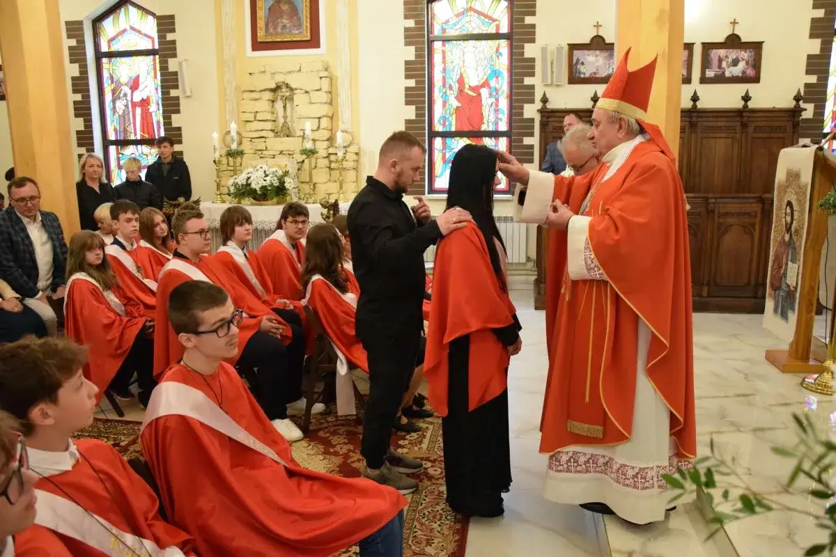Biskup udziela sakramentu bierzmowania. Ceremonia trwa, a młodzież w czerwonych szatach czeka na swoją kolej.