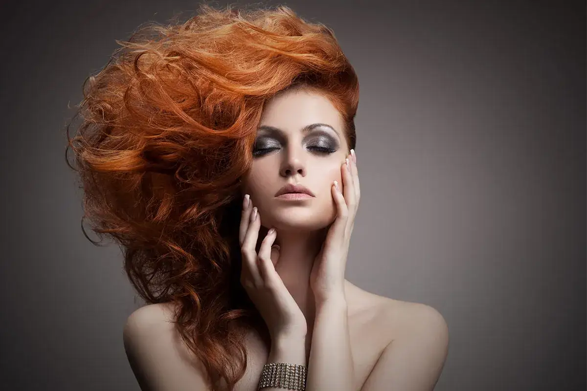 Femme aux cheveux roux flamboyants, maquillage intense, mains sur le visage. Le vignettage photo accentue le mystère.