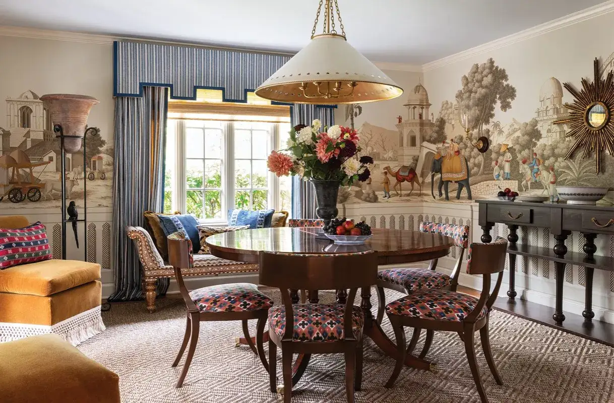 A round dining table with patterned chairs showcases eclectic table styles. The room features a mural of an Indian landscape, striped curtains, and plush seating.