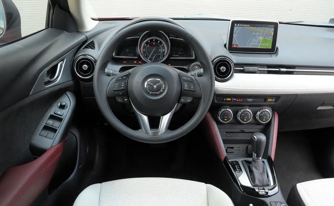Wnętrze Mazdy CX-3: kierownica, nawigacja, panel klimatyzacji i dźwignia zmiany biegów. Atrakcyjna mazda cx3 cena zachęca do zakupu.