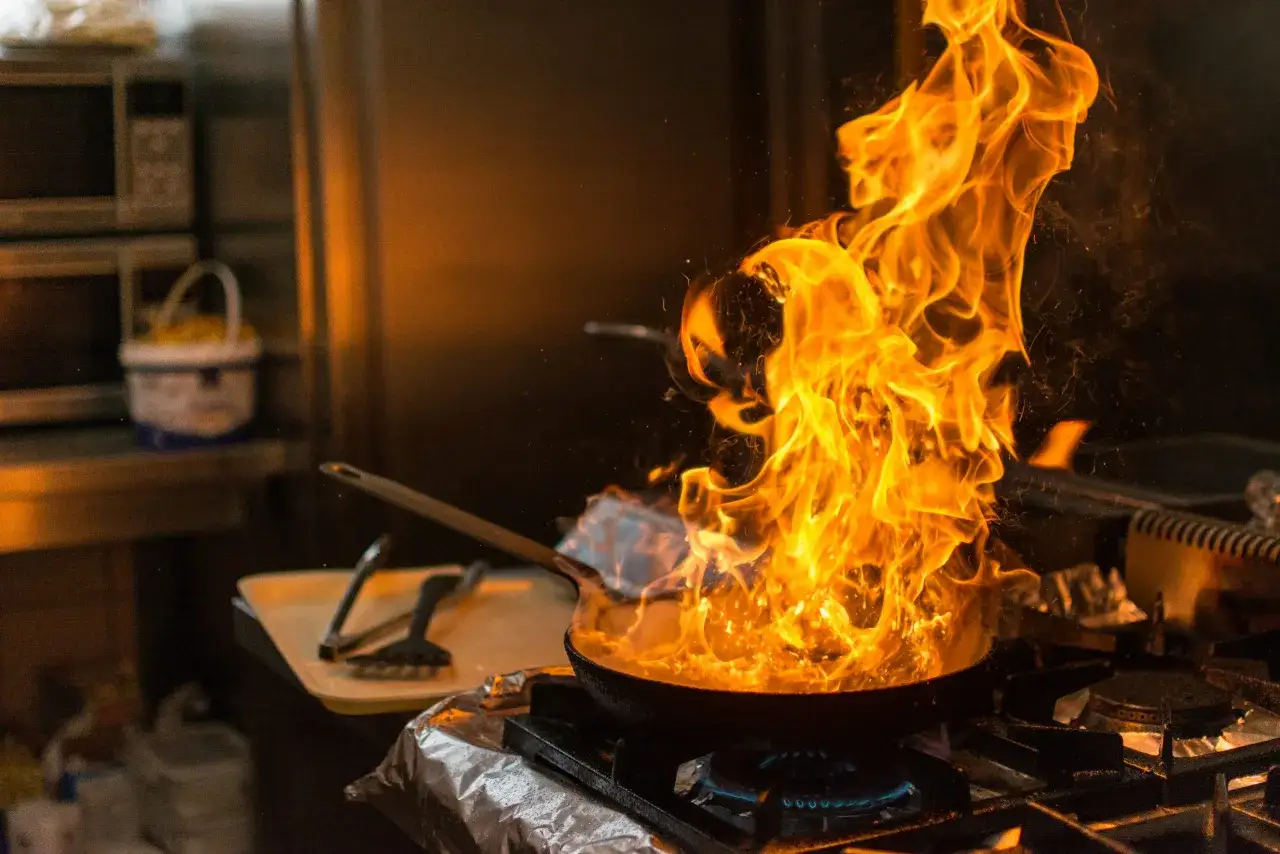 Eine brennende Pfanne auf dem Herd, Flammen schlagen hoch. Ein Koch bereitet ein Gericht zu.