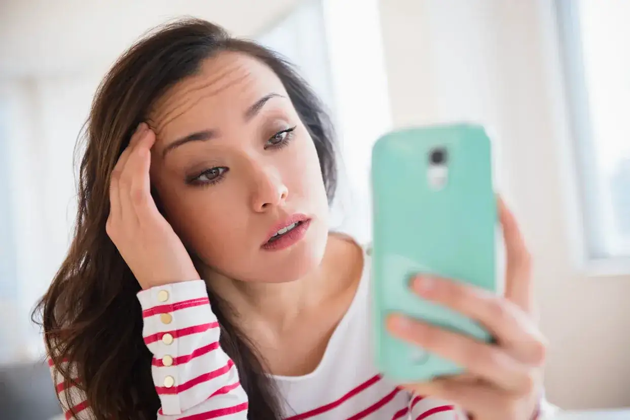 Een vrouw met fronsende wenkbrauwen kijkt naar haar telefoon, haar hand aan haar voorhoofd. Ze lijkt verslaafd aan prikkels, verloren in de digitale wereld.