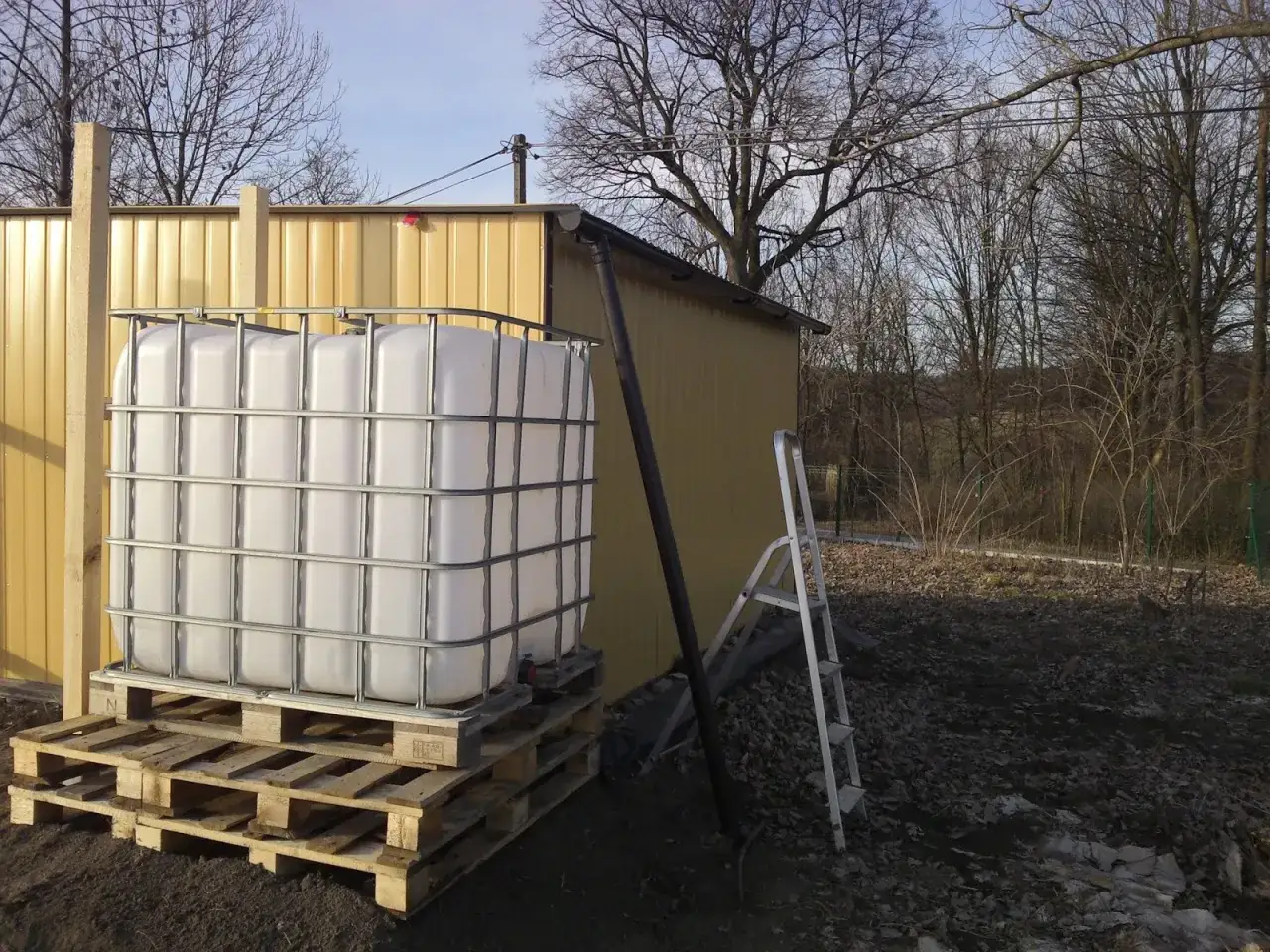 Zbiornik IBC na paletach obok żółtej szopy, z rynną prowadzącą do ziemi. W tle drzewa i drabina. Jak podłączyć mauser do rynny?