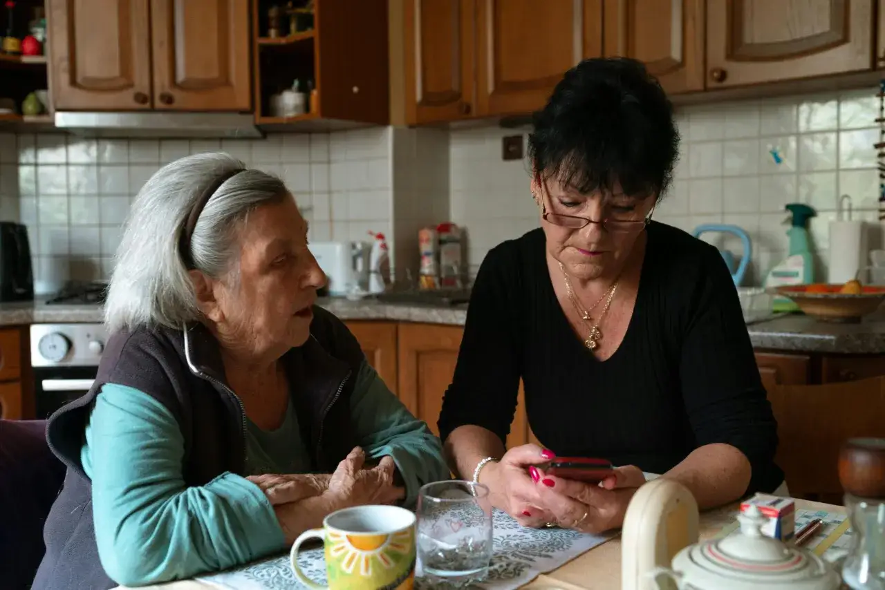 Rozmowa o tym, ile pieniędzy za opiekę nad starszą osobą, przy kawie. Kobieta z siwymi włosami mówi, druga patrzy w telefon.