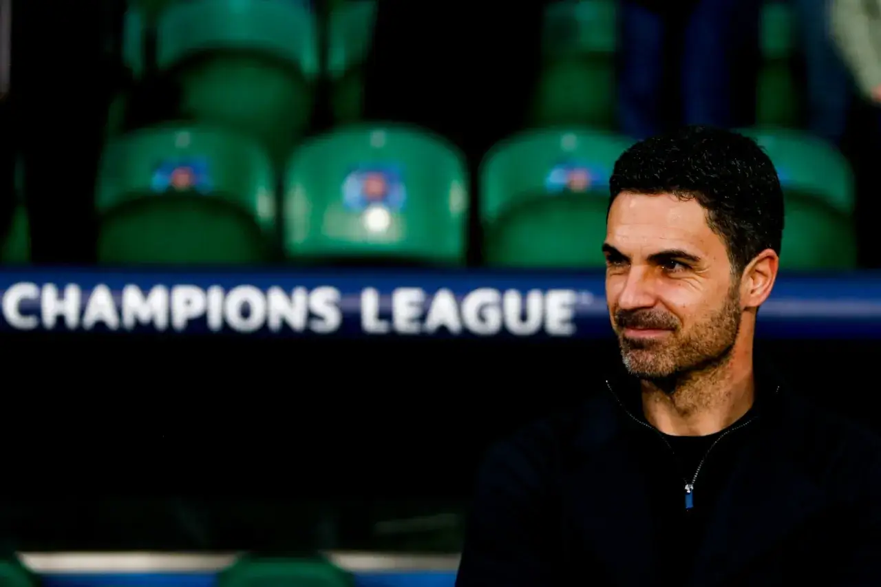Mikel Arteta, trener Arsenalu, z uśmiechem patrzy w bok. W tle widać zielone siedzenia i napis