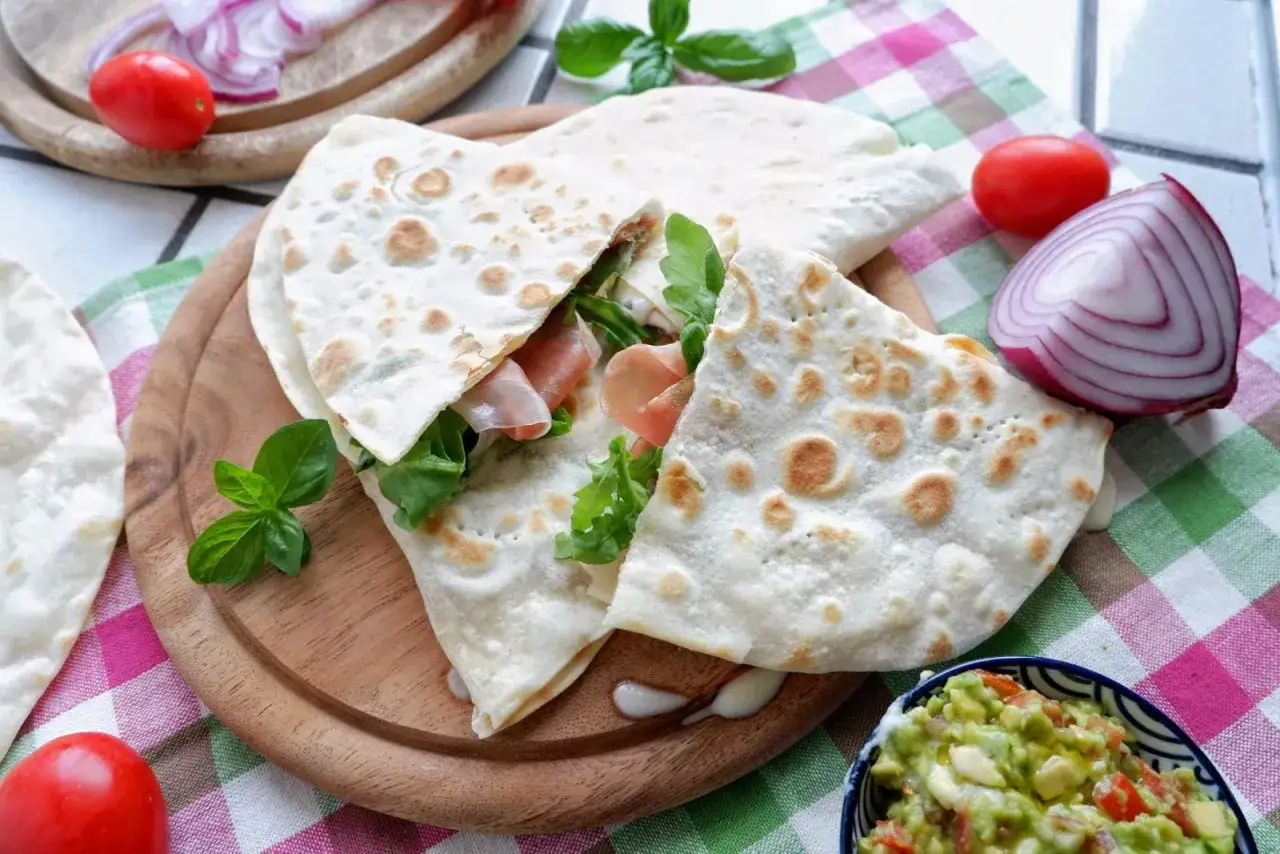Piadina fatta in casa ripiena di prosciutto e rucola, servita con guacamole e ingredienti freschi.