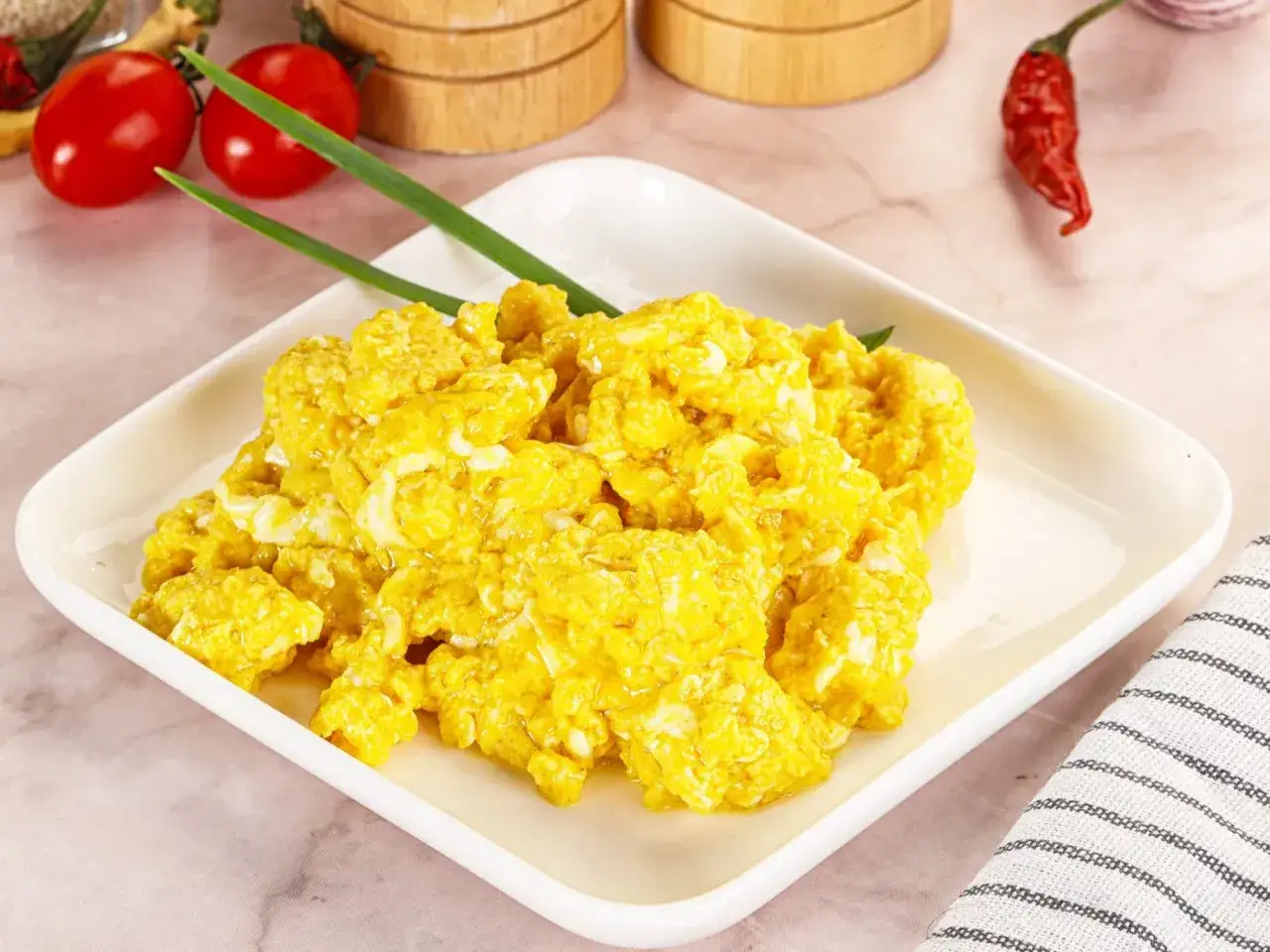 Puszysta jajecznica z serem, udekorowana szczypiorkiem, na białym talerzu. Obok pomidorki i papryczka chili.