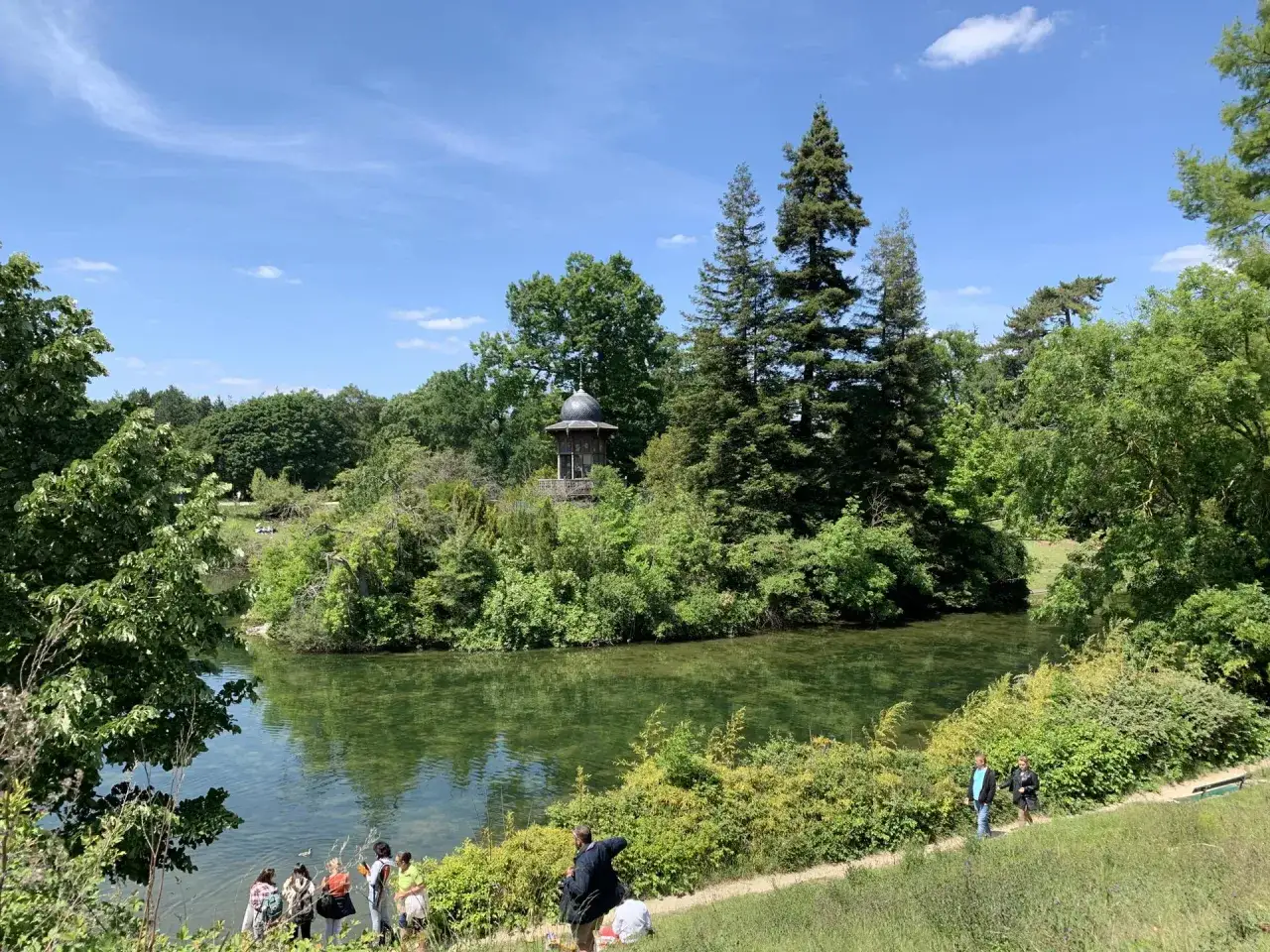 Un d&iacute;a soleado en el Bois de Boulogne, con un lago sereno, vegetaci&oacute;n exuberante y una torre con c&uacute;pula.