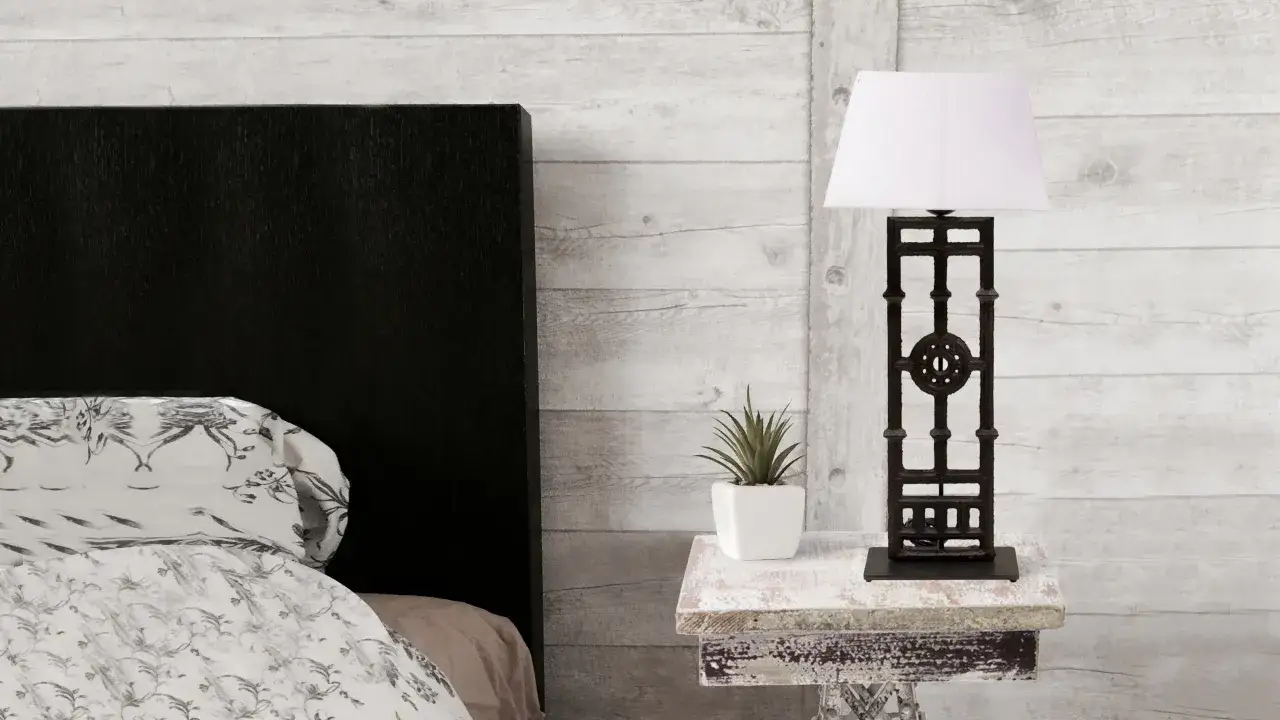 Lámpara de forja de techo con pantalla blanca sobre mesita auxiliar, junto a una cama con cabecero negro y ropa de cama estampada.