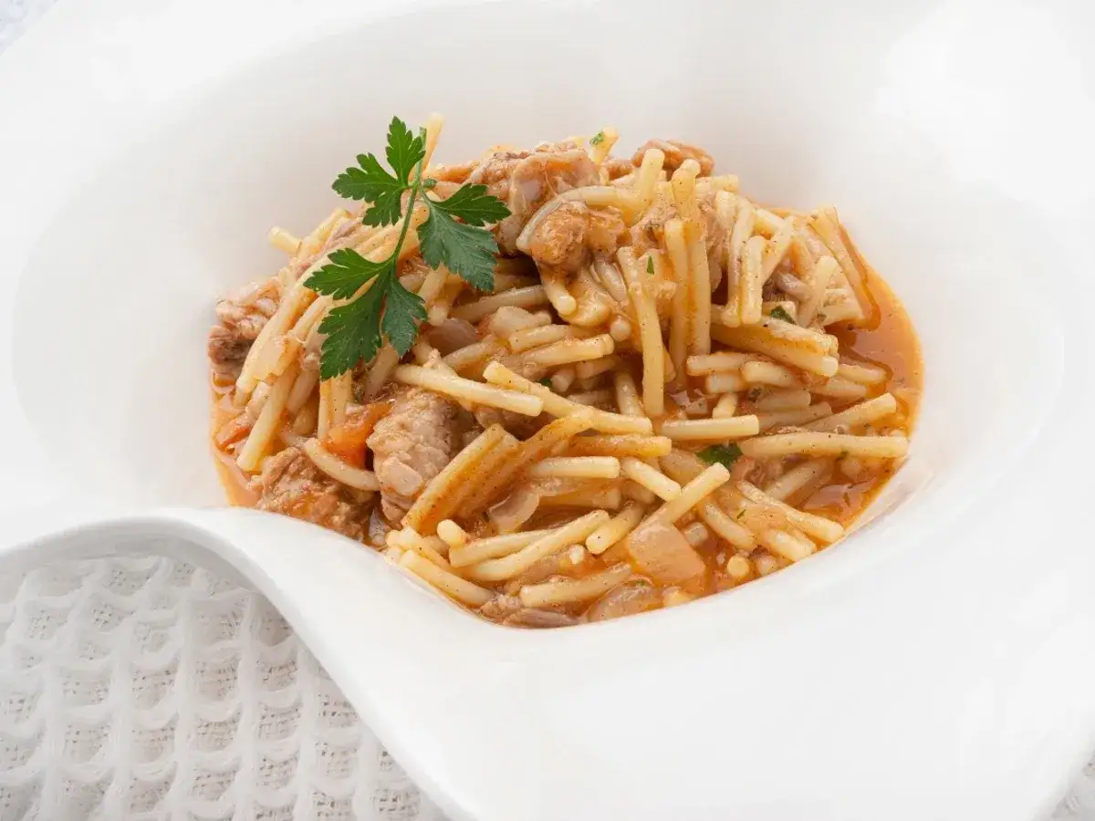 Un plato de fideos a la cazuela con trozos de carne y perejil fresco, servido en un cuenco blanco.