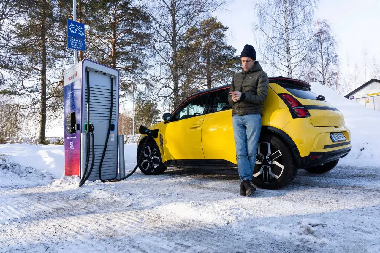 Man kollar mobilen medan en gul elbil laddas i snön. Ett elbil test i vinterväglag.