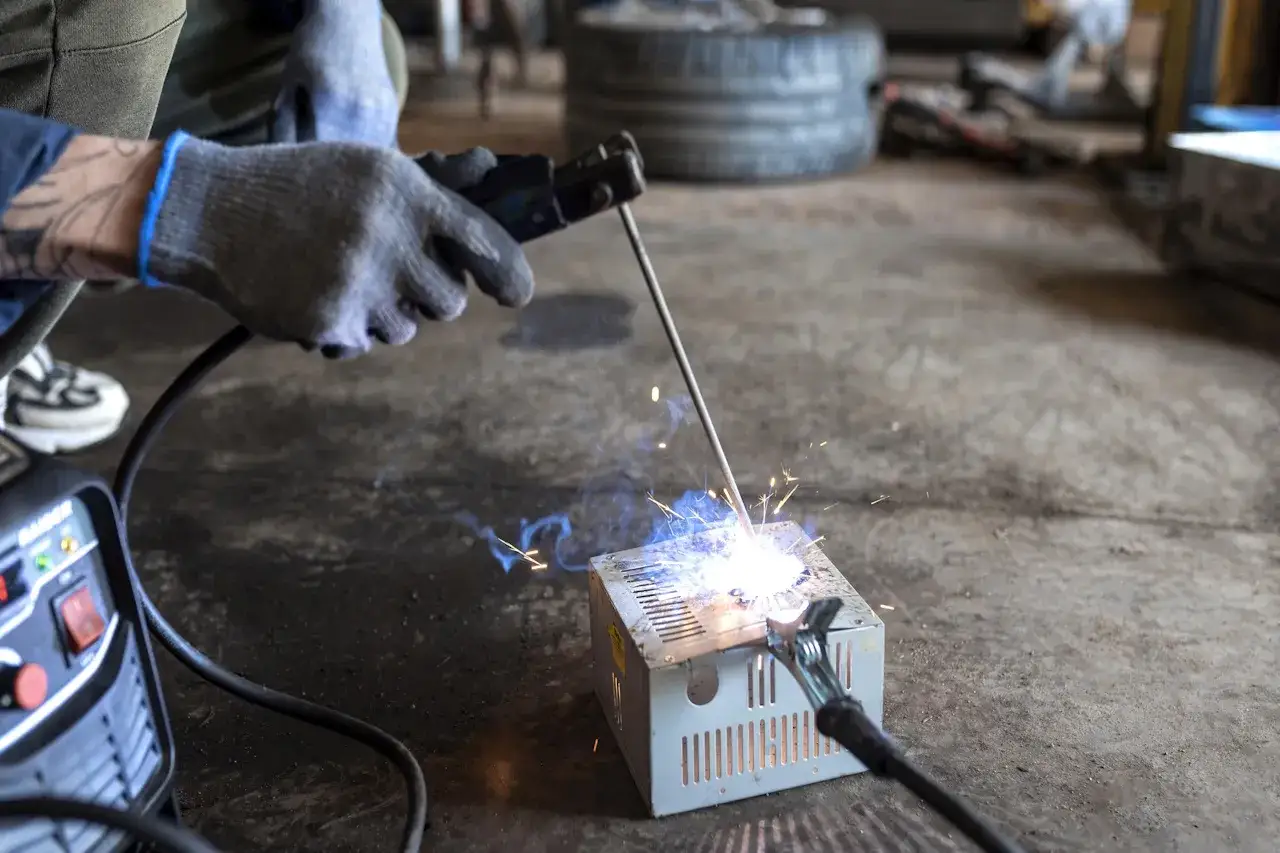 Ręka w rękawicy spawa migomatem aluminiową obudowę. Iskry i niebieski łuk spawalniczy oświetlają warsztat.