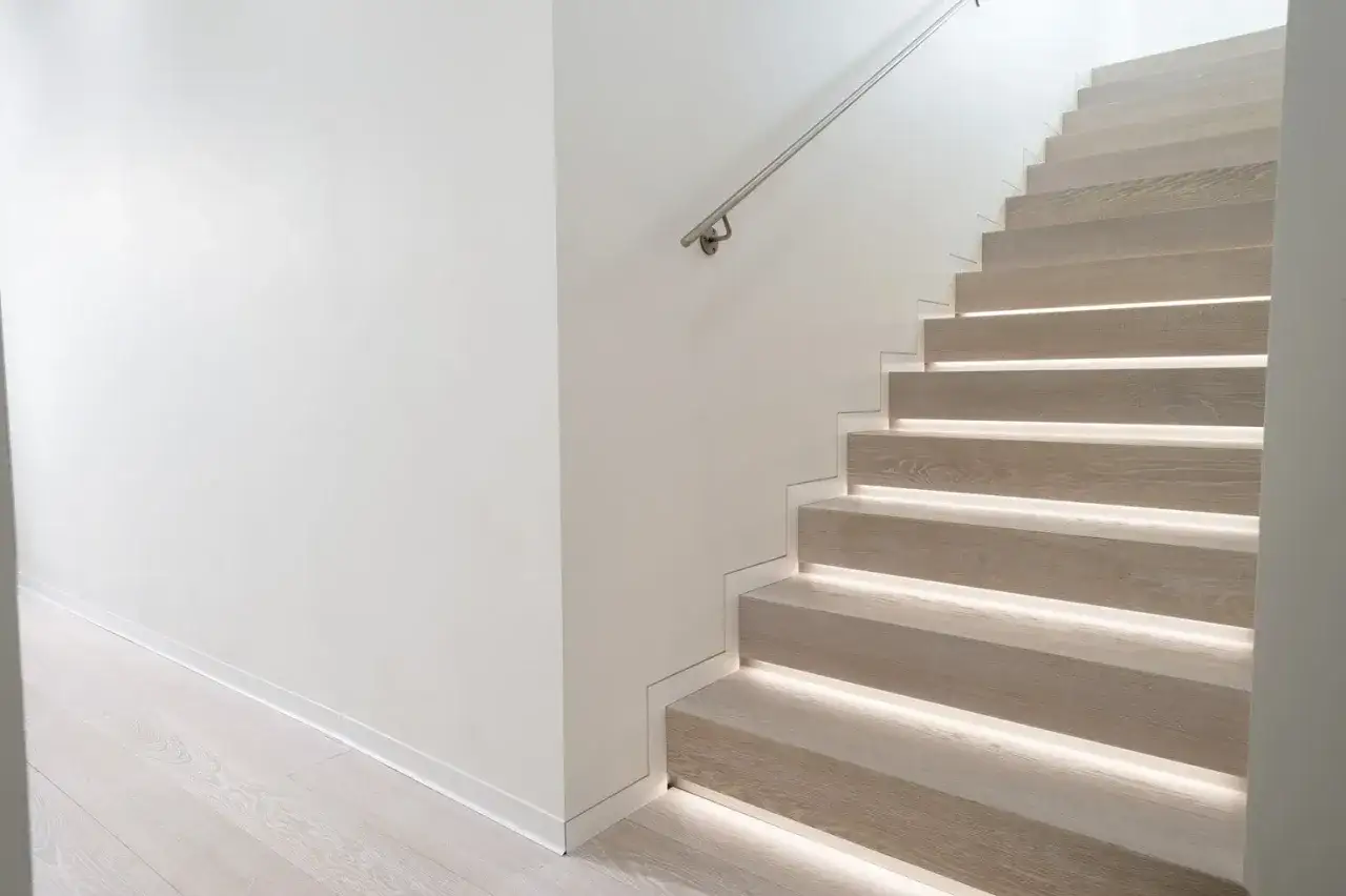 Scala moderna con gradini in legno illuminati. Il battiscopa filo muro crea un effetto pulito, ma valuta pro e contro per la tua casa.