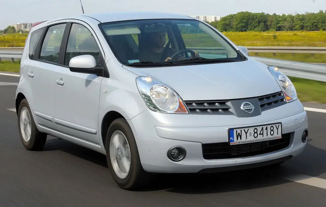 Srebrny Nissan Note 2010 jedzie po drodze, za kierownicą siedzi mężczyzna.