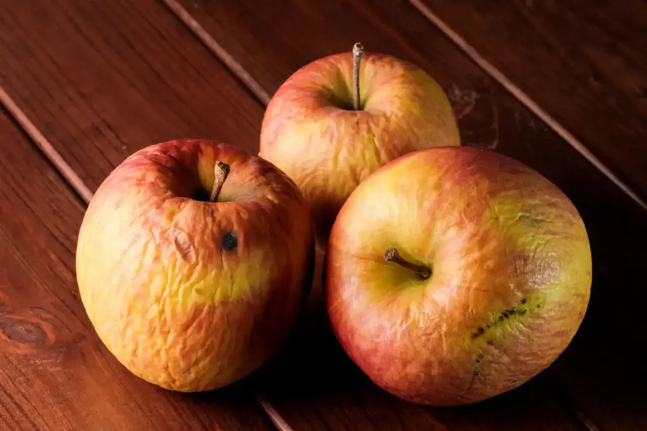 Trois pommes légèrement ridées sur une table en bois. Parfaites pour savoir que faire avec des pommes abîmées : compote, tarte ou jus !