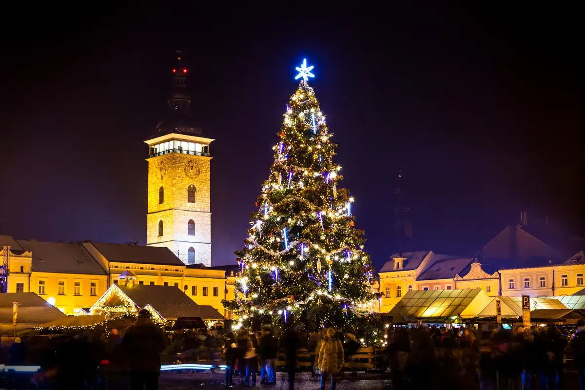 Festlicher budweis weihnachtsmarkt mit beleuchtetem Baum und historischem Turm. Menschen schlendern zwischen den St&auml;nden.
