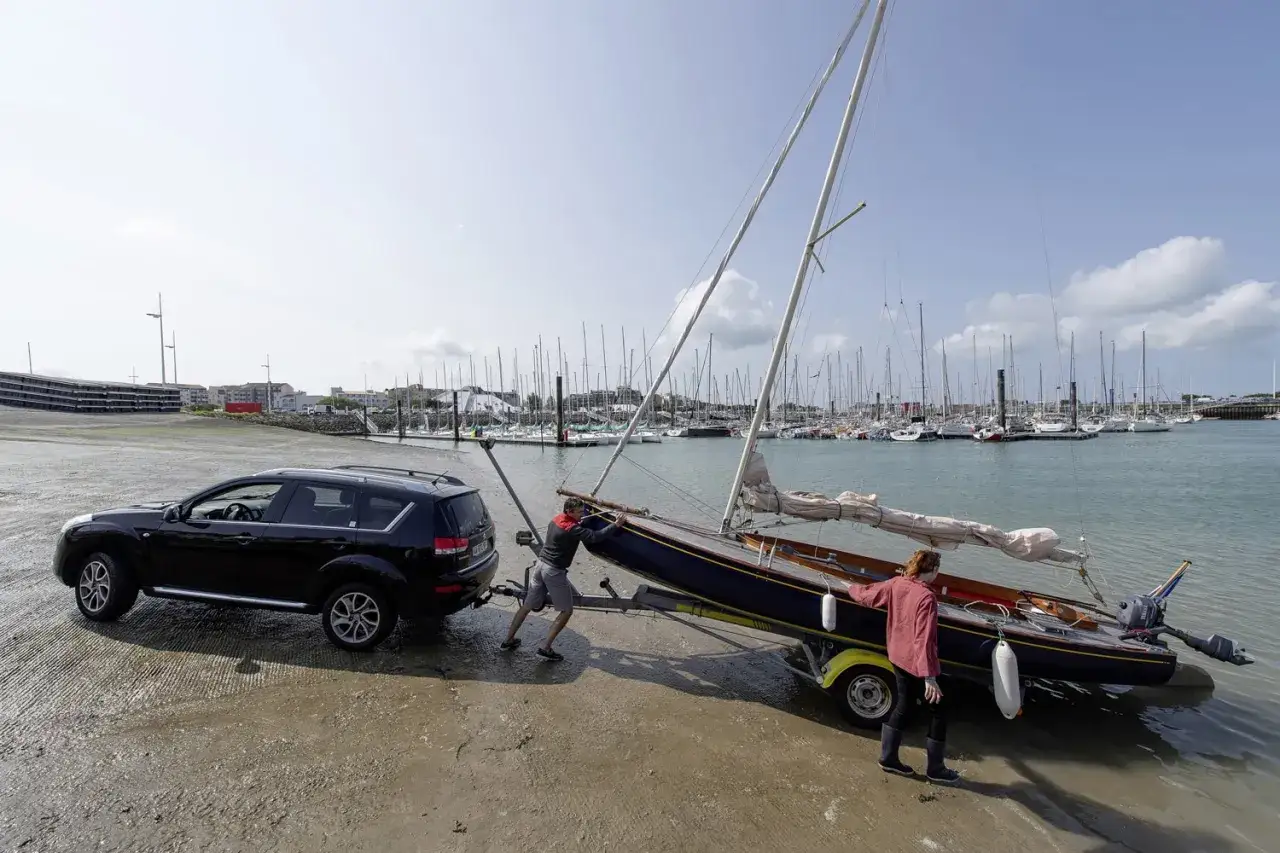 Deux personnes aident à mettre à l'eau un voilier sur une remorque tractée par une voiture. Le port est rempli de bateaux. Heol 7.4.