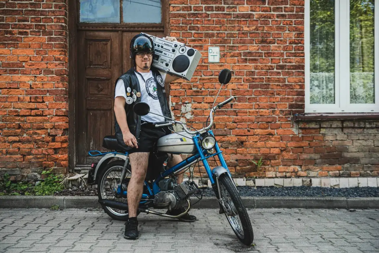 Młody mężczyzna w kasku i kamizelce, z głośnikiem na ramieniu, siedzi na motorowerze Romet. Wygląda jak prawdziwy ogar gotowy na muzyczną podróż.