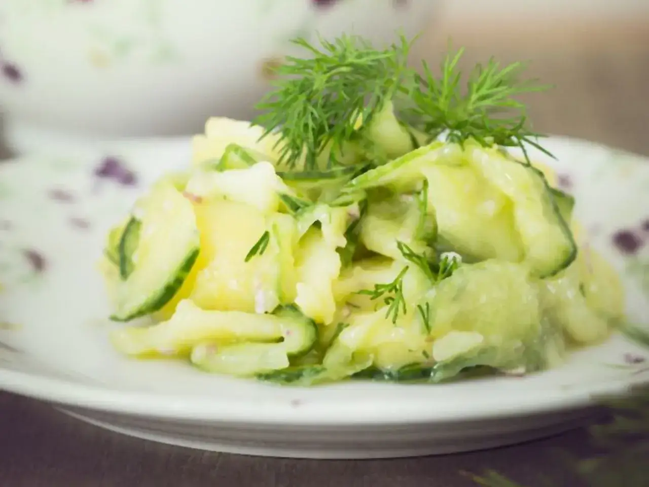 Ein cremiger Kartoffelsalat mit Gurken und frischem Dill, serviert auf einem Teller mit zartem Blumenmuster.
