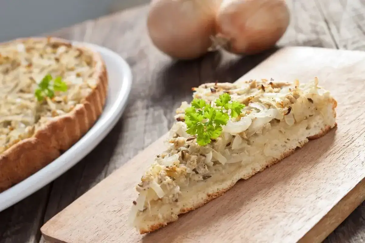 Une part de tarte à l'oignon, garnie de persil frais, repose sur une planche en bois. Une tarte entière est visible sur une assiette blanche à gauche.
