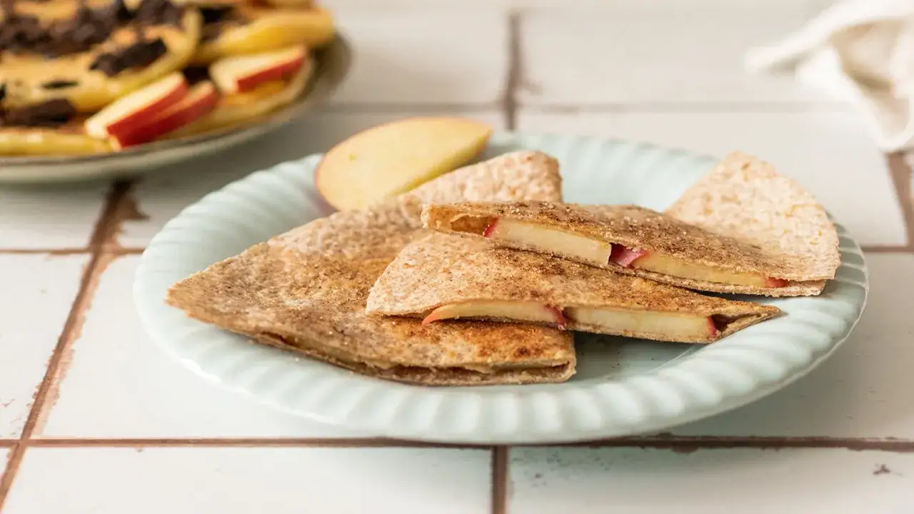 Słodka tortilla z jabłkami i cynamonem, pokrojona na tr&oacute;jkąty, leży na talerzu. Obok widać plasterki jabłka.