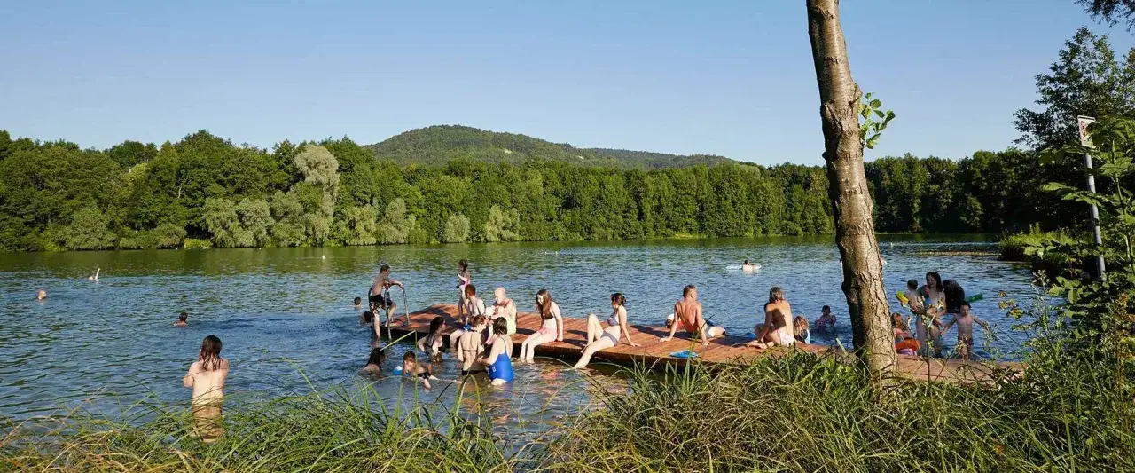 Menschen genie&szlig;en einen sonnigen Tag am baggersee Happurg. Sie sitzen auf einem Holzsteg, schwimmen und entspannen sich im Wasser.