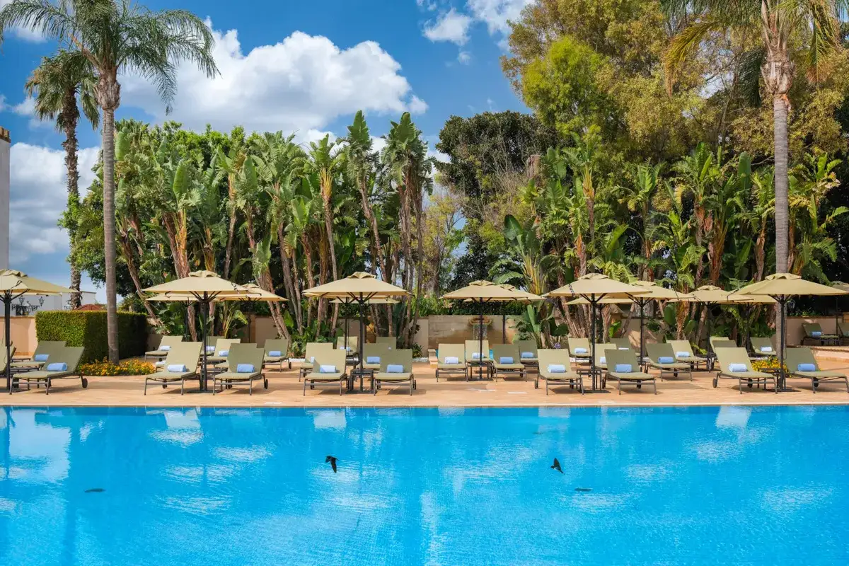 Piscina azzurra circondata da lussureggianti giardini siciliani, lettini e ombrelloni invitano al relax sotto il sole.