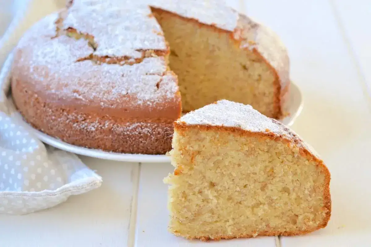 Una fetta di torta di banane mature, soffice e cosparsa di zucchero a velo, accanto alla torta intera su un piatto bianco.