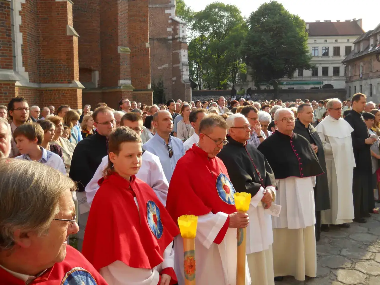 Kanonik w czarnej sutannie stoi w tłumie wiernych przed ceglanym kościołem. W ręku trzyma zapaloną świecę.