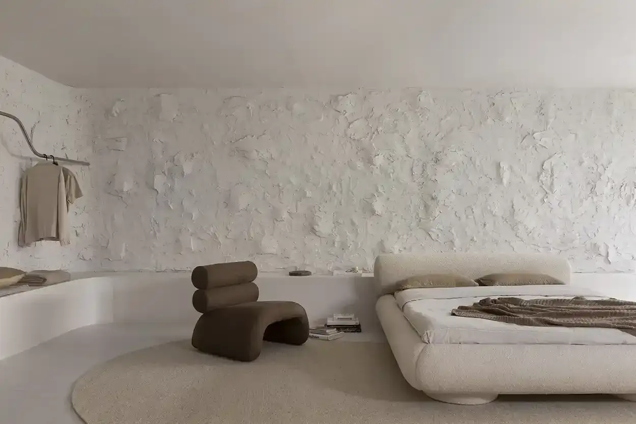 Habitación minimalista con cama baja, sillón curvo y pared con textura de gotele moderno en blanco roto.