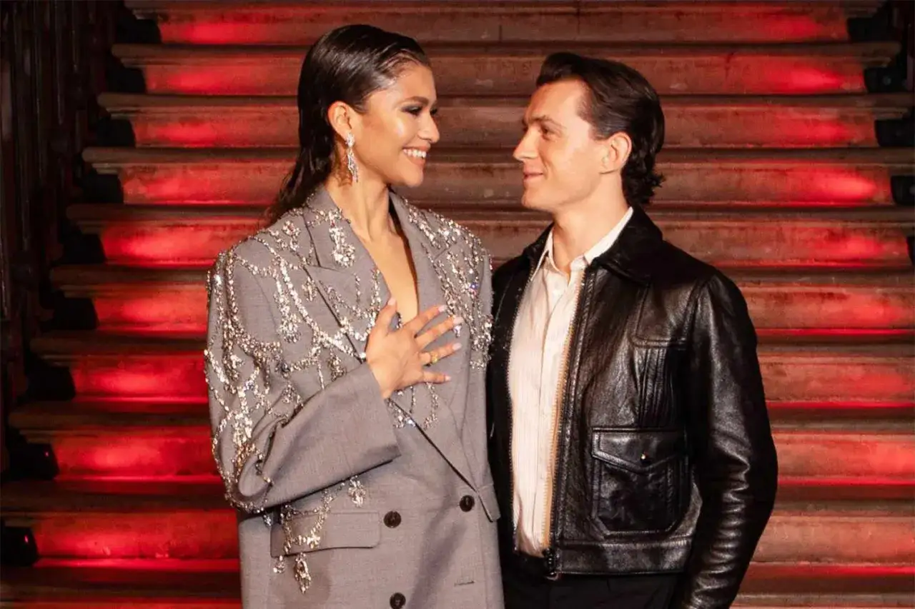 Tom Holland i Zendaya, uśmiechnięci, stoją na tle czerwonych schodów. Ona w błyszczącej marynarce, on w skórzanej kurtce.