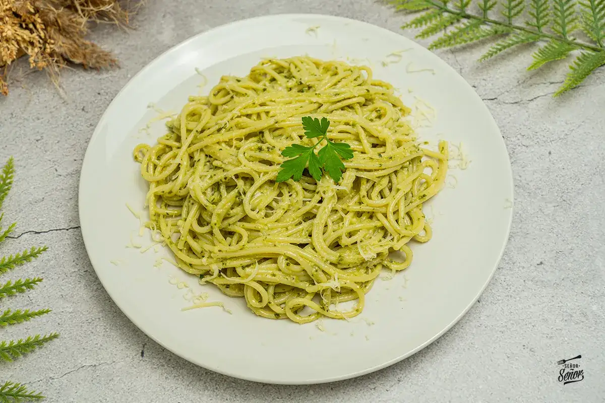 Un plato de pasta al pesto, con espaguetis cubiertos de salsa verde y queso rallado, decorado con una ramita de perejil.